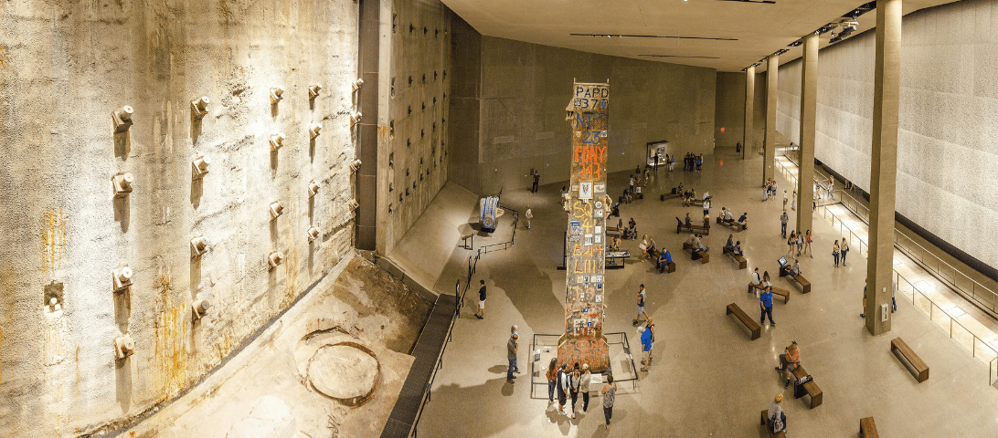 Panoramic view of the 9/11 Memorial Museum showing exhibit items and visitors. Central is a large artifact display surrounded by people. The background features the museum's concrete walls and other exhibits, making it one of the essential things to do when in New York City.