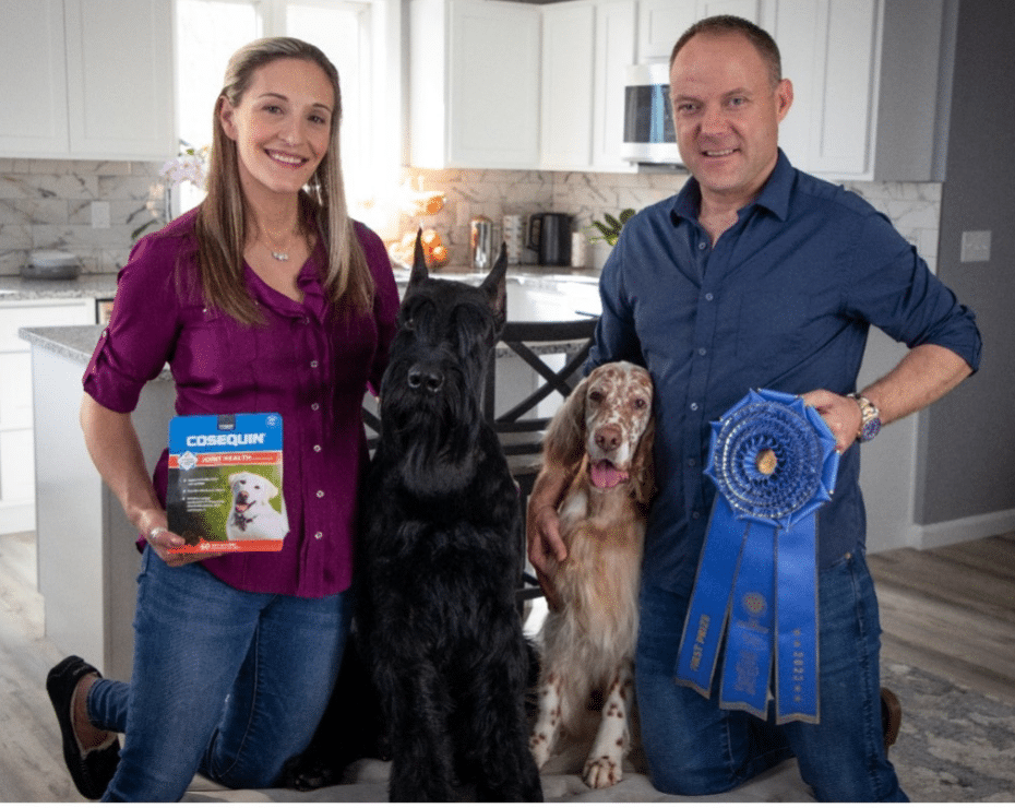 A woman and a man are kneeling in a kitchen with two large dogs, holding a box of Cosequin and a blue ribbon, respectively.