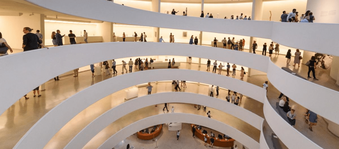 A multi-level spiral gallery offers numerous things to do, with people viewing art exhibits on each floor, featuring white curved walls and a central open atrium.