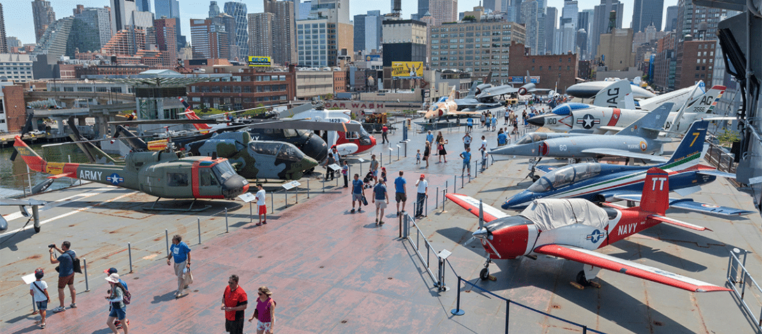 Visitors seeking engaging things to do can observe various military aircraft displayed outdoors on a ship deck, with a city skyline in the background.