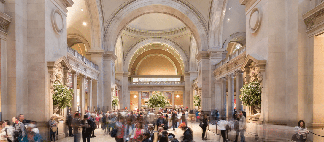 A bustling crowd inside a large hall with arched ceilings and ornate architecture. The hall is illuminated with soft lighting, and potted plants are placed around the area, making it one of the exciting things to do for those who appreciate architectural beauty.