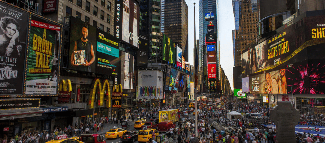 A busy urban street scene in Times Square, New York City, with numerous advertisements, neon signs, and crowds of people. Yellow taxis are visible amidst the pedestrian-filled area, making it one of the top things to do for any visitor exploring the city's vibrant atmosphere.