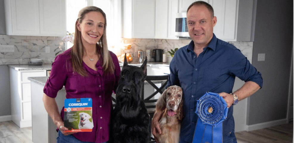 In a cozy kitchen scene fit for a blog post, a woman holds a pet supplement box while the man beside her proudly displays a blue ribbon. Between them sit two dogs, one black and the other brown with white, completing the heartwarming picture.