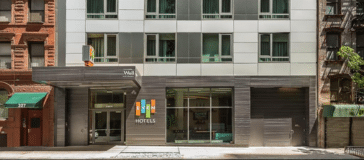 Exterior of a modern hotel building with a sign that reads "Hotels" near the entrance. The facade features large windows and a combination of metal and brick elements, showcasing sleek design and contemporary architecture.
