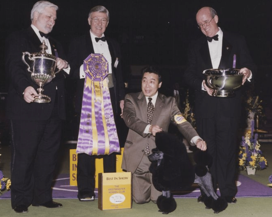 Four men in formal attire stand on a stage. One holds a large ribbon, another holds a trophy, and the third holds a large silver bowl. The fourth man kneels beside a black dog.