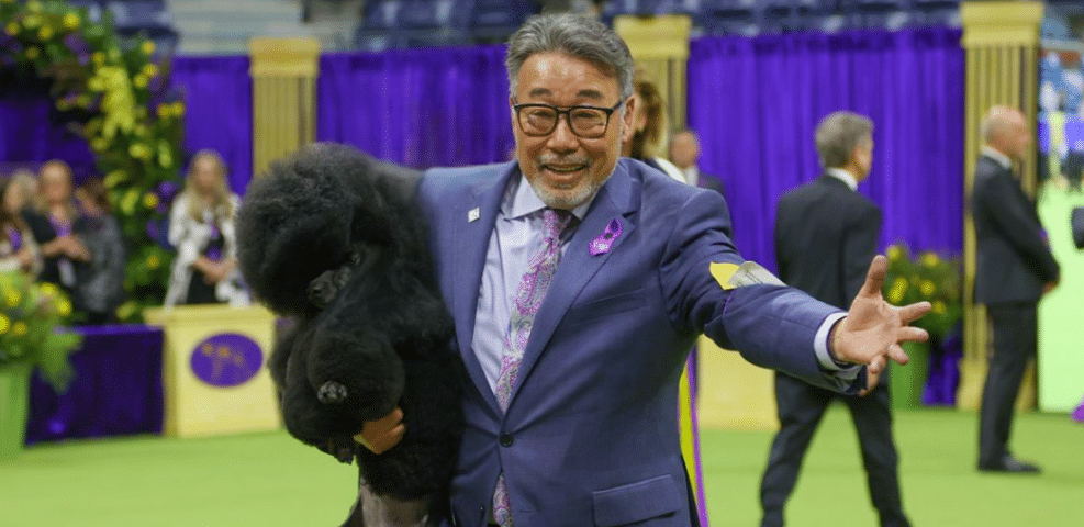 A man in a suit holds a black poodle in one arm and gestures with his other hand while standing in an indoor arena, decorated with purple and yellow elements, as though he's about to share his story on his blog.