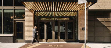 Entrance of a modern building with glass doors, brick facade, and a canopy decorated with small lights. A person walks past the building, resembling those grand hotels in the city. A sign is posted on the sidewalk nearby.