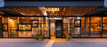 Exterior of a modern building with large glass windows, decorative lighting on the overhang, and potted plants beside the entrance. The windows display the words "The Vine" and "851 Woodmoor," giving it an inviting hotel-like ambiance.