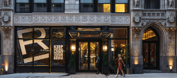 A street view of a modern boutique hotel entrance with a large "R" on the window and the text "KEEP ROLLING WITH US, ROCKY START? PEEN!" Hotels can add charm to any journey. Two people are walking past the entrance.