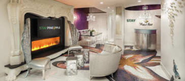 Modern hotel lobby with a fireplace, mirrored fire sculpture, plush chairs, chandelier lighting, and a reception desk. The words "STAY PINEAPPLE" are displayed prominently, showcasing the unique charm found in the best hotels.