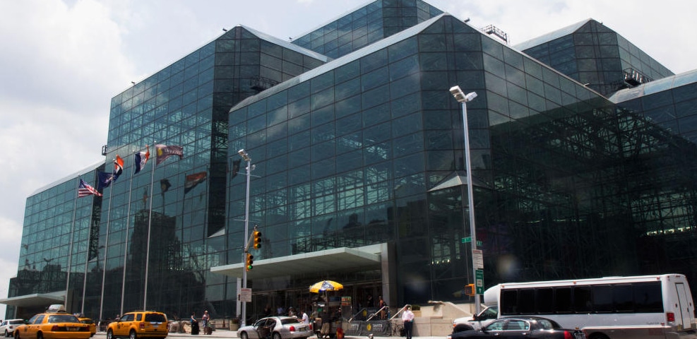 A large modern glass building, bustling with exhibitors, stands proudly with multiple flags in front. Several vehicles are parked nearby under the watchful gaze of a cloudy sky above.