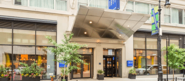 Street-level view of a modern hotel building with a glass entrance, an overhang, large windows, and potted plants on either side of the entryway.