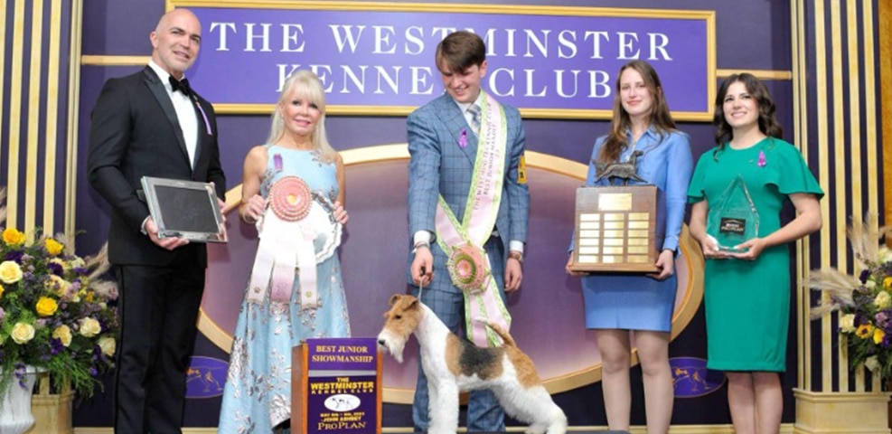 A group of five people and a dog pose for a blog-worthy photo at the Westminster Kennel Club event, proudly holding awards and ribbons.