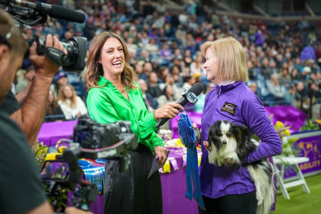 A woman in a bright green shirt interviews another woman holding a dog and two blue ribbons at a dog show, surrounded by cameras and an audience. It's the perfect scene for highlighting holiday gifts for dog lovers in this festive and joyful atmosphere.