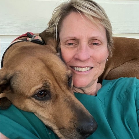 A person smiles while cuddling with a large brown dog resting its head on their shoulder, embodying the compassion and dedication worthy of Veterinarian of the Year.